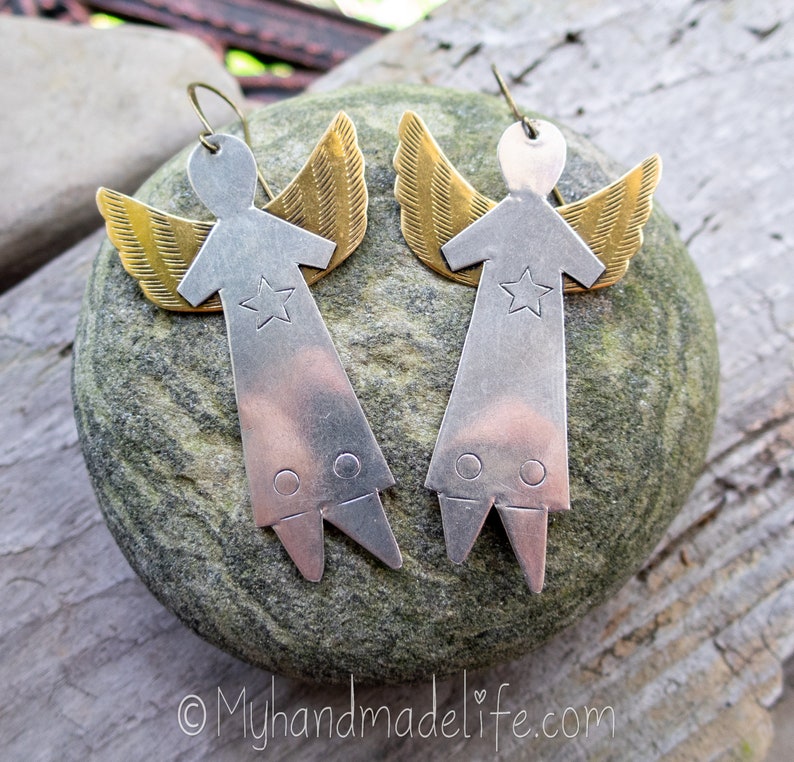 May include: A pair of handmade angel earrings. Each earring has a silver-coloured body with a star and gold-coloured wings. The earrings are displayed on a grey stone, with a wooden background. The earrings have a hook closure.