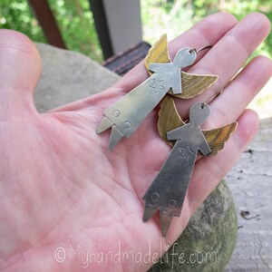 May include: A pair of handmade angel earrings. Each earring has a silver-coloured body with a star and brass-coloured wings. The earrings are held in a hand, highlighting their design and craftsmanship. The earrings have a unique, artistic style.