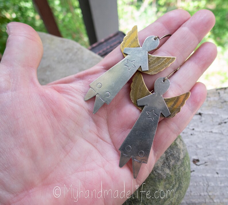 May include: A pair of handmade angel earrings. Each earring has a silver-coloured body with a star and brass-coloured wings. The earrings are held in a hand, highlighting their design and craftsmanship. The earrings have a unique, artistic style.