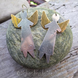 May include: A pair of handmade angel earrings. Each earring has a silver-coloured body with a star and two circles, and gold-coloured wings. The earrings are displayed on a grey stone.
