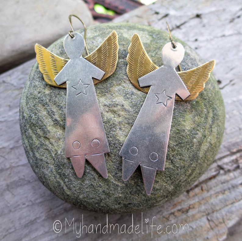May include: A pair of handmade angel earrings. Each earring has a silver-coloured body with a star and two circles, and gold-coloured wings. The earrings are displayed on a grey stone.