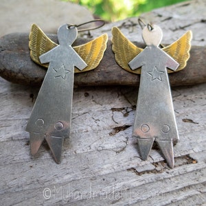 May include: A pair of handmade angel earrings. Each earring has a silver-coloured body with a star and two circles, and gold-coloured wings. The earrings are displayed on a wooden surface.