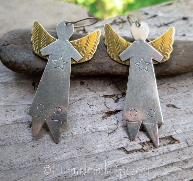 May include: A pair of handmade angel earrings. Each earring has a silver-coloured body with a star and two circles, and gold-coloured wings. The earrings are displayed on a wooden surface.