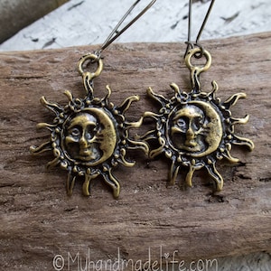 Könnte beinhalten: Antike bronzefarbene Ohrringe mit einem Sonnen- und Mond-Design. Jeder Ohrring zeigt ein detailliertes Gesicht innerhalb des Mondes, umgeben von Sonnenstrahlen. Die Ohrringe hängen an einfachen Drahtbügeln. Der Hintergrund ist eine verwitterte Holzoberfläche.