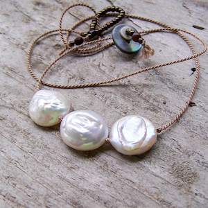 Peut inclure: Un collier fait à la main avec trois grandes perles baroques blanches iridescentes. Les perles sont enfilées sur un cordon brun clair délicat, avec un fermoir décoratif et des perles. Le collier est présenté sur une surface en bois vieilli.