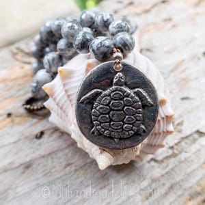 May include: A necklace with grey and white speckled beads and a large, round, silver-toned pendant with a detailed turtle design. The necklace rests on a white seashell, with the text "Myhandmadelife.com" in the bottom corner.