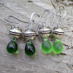 May include: Four pairs of frog earrings. Each earring has a silver-coloured frog head and a dark green or light green teardrop-shaped glass bead. The earrings hang from silver-coloured ear wires. The background is a textured wooden surface.