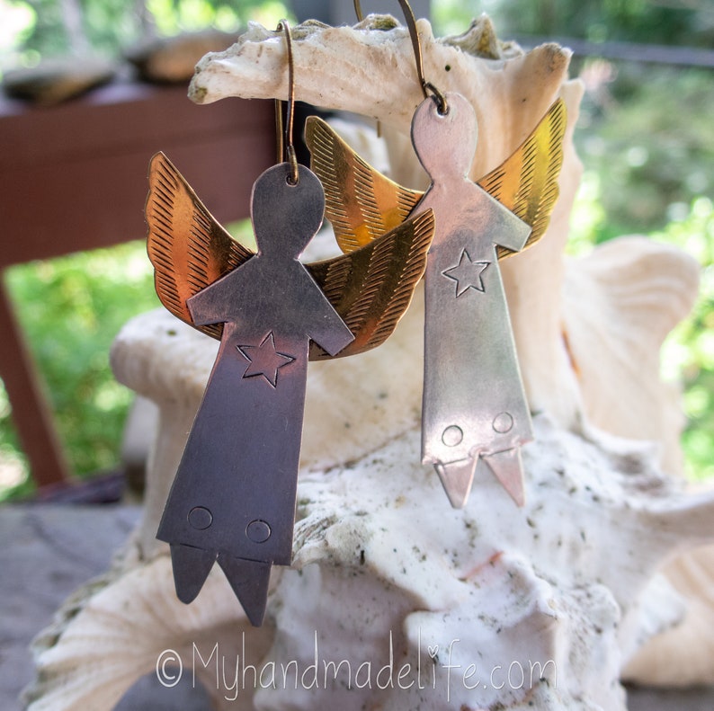May include: Handmade angel earrings. Each earring features a metal angel figure with a star cutout and detailed wings. The earrings are displayed against a natural, textured background, showcasing their handcrafted nature.