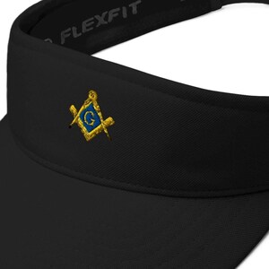 Pode incluir: Viseira Flexfit preta com um emblema dourado e azul. O emblema apresenta um design de esquadro e compasso com a letra "G" no centro. A viseira tem uma alça ajustável para um ajuste confortável.