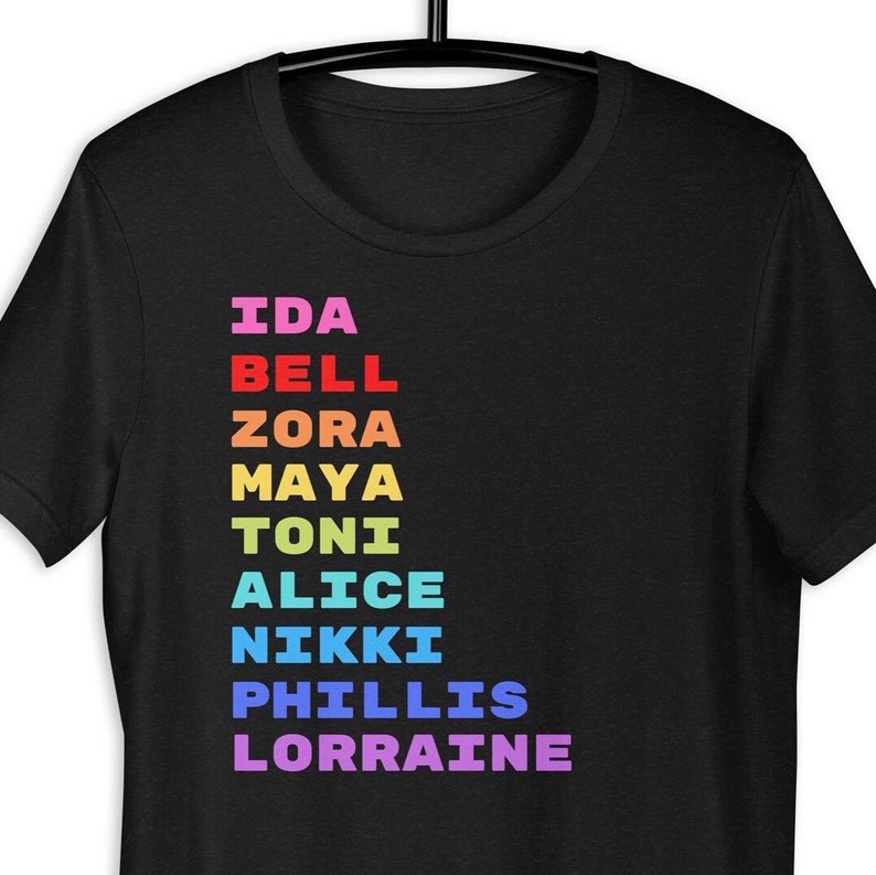 K&ouml;nnte beinhalten: Schwarzes T-Shirt mit einer senkrechten Liste von Namen in Regenbogenfarben: Ida Bell, Zora, Maya, Toni, Alice, Nikki, Phillis, Lorraine.