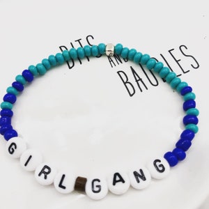 K&ouml;nnte beinhalten: Ein Armband mit den Worten "GIRL GANG" in wei&szlig;en Buchstabenperlen. Das Armband hat blaue und t&uuml;rkisfarbene Perlen sowie einen kleinen silbernen Akzent. Das Armband liegt auf einer wei&szlig;en Oberfl&auml;che.