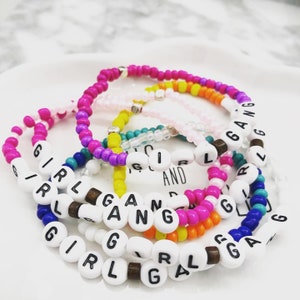 K&ouml;nnte beinhalten: Eine Sammlung farbenfroher Perlenarmb&auml;nder. Jedes Armband hat wei&szlig;e Buchstabenperlen mit der Aufschrift "GIRL GANG". Die Armb&auml;nder sind in den Farbt&ouml;nen Rosa, Orange, Gelb, Blau und Wei&szlig; gehalten, mit Holz- und Silberakzenten.