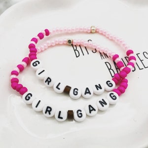 K&ouml;nnte beinhalten: Zwei Armb&auml;nder mit Perlen. Eines ist rosa und wei&szlig;, das andere ist rosa mit wei&szlig;en Buchstabenperlen, die "GIRL GANG" buchstabieren. Die Armb&auml;nder liegen auf einem wei&szlig;en Teller.