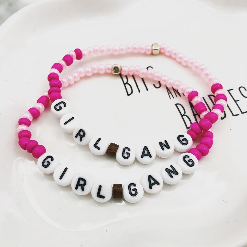 K&ouml;nnte beinhalten: Zwei Armb&auml;nder mit Perlen. Eines ist rosa und wei&szlig;, das andere ist rosa mit wei&szlig;en Buchstabenperlen, die "GIRL GANG" buchstabieren. Die Armb&auml;nder liegen auf einem wei&szlig;en Teller.