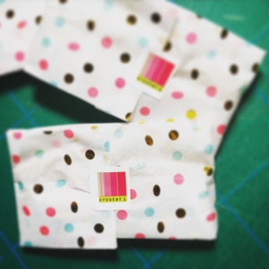 K&ouml;nnte beinhalten: Mehrere Pakete, verpackt in wei&szlig;em Papier mit Polka-Dots mit rosa, blauen, gelben und braunen Punkten. Jedes Paket hat einen Aufkleber mit dem Text "crystal j." in einem rosa-goldenen Design.