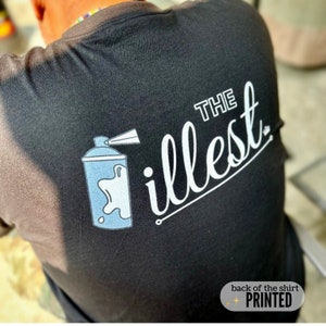 May include: Black t-shirt with white text that says "THE illest" and a blue spray paint can graphic. The text is in a cursive font. The back of the shirt is printed.