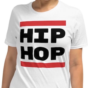May include: White t-shirt featuring the words "HIP HOP" in large, bold, black letters. The text is centered and framed by a thin red horizontal stripe above and below. The shirt is made of a soft material.