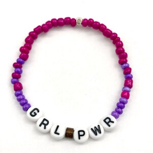 May include: A pink and purple beaded bracelet with the word "GRL PWR" spelled out in white beads.