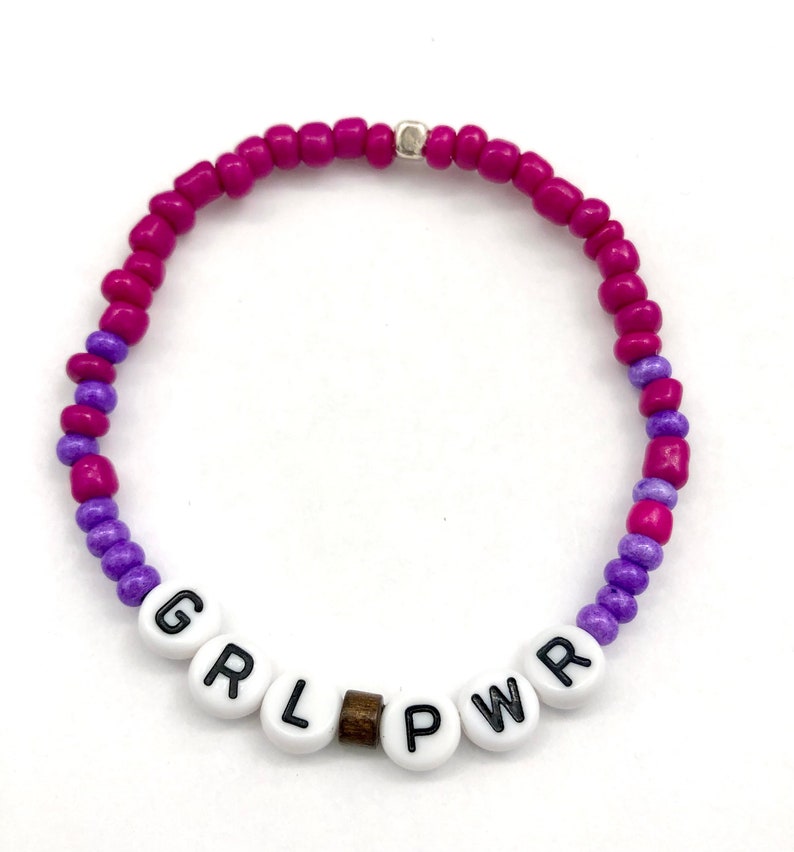 May include: A pink and purple beaded bracelet with the word "GRL PWR" spelled out in white beads.