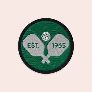 May include: A green and white embroidered patch with two crossed paddleball rackets and a ball in the center. The text "EST. 1965" is embroidered on the rackets.