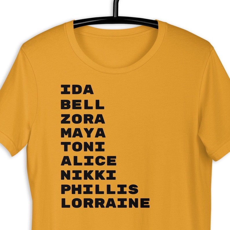 K&ouml;nnte beinhalten: Ein gelbes T-Shirt mit schwarzem Text, der die Namen Ida Bell, Zora, Maya, Toni, Alice, Nikki, Phillis und Lorraine auflistet.