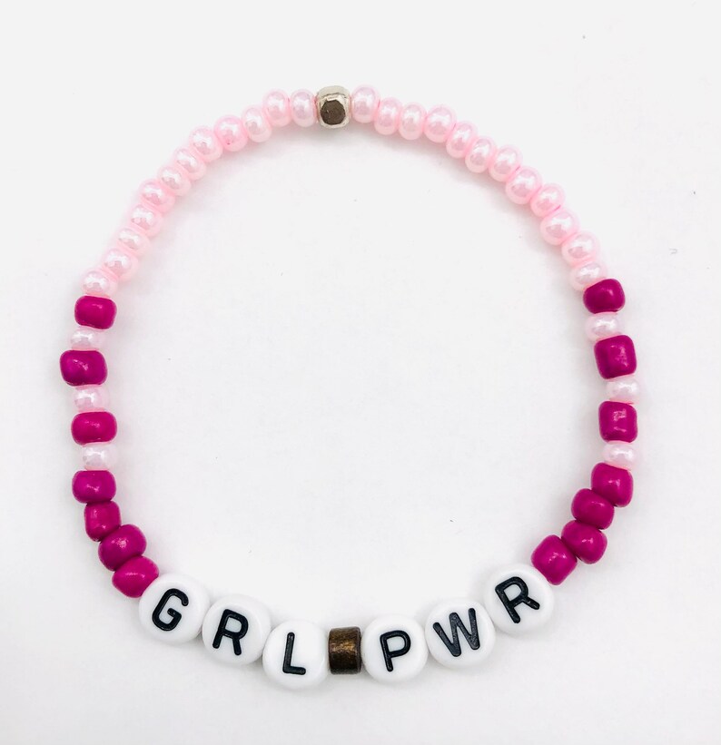 May include: Pink and magenta beaded bracelet with the word "GRL PWR" spelled out in white letters.