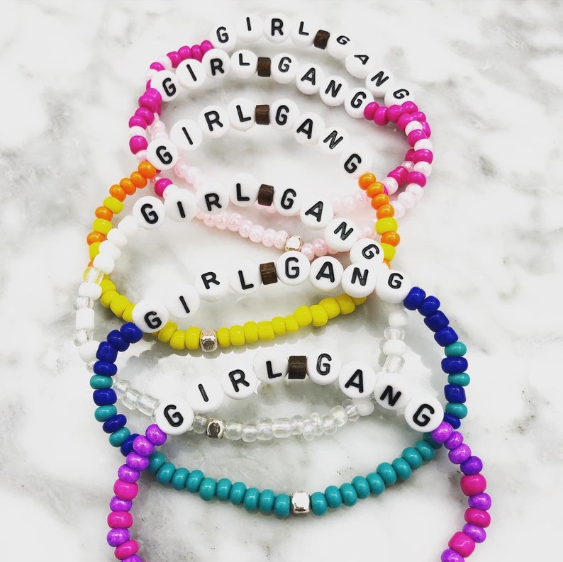 K&ouml;nnte beinhalten: Eine Reihe von Armb&auml;ndern aus Perlen in verschiedenen Farben, darunter Rosa, Gelb, Blau und T&uuml;rkis. Jedes Armband schreibt "GIRL GANG" mit wei&szlig;en Buchstabenperlen und Holzperlen. Die Armb&auml;nder liegen auf einer wei&szlig;en Marmoroberfl&auml;che.