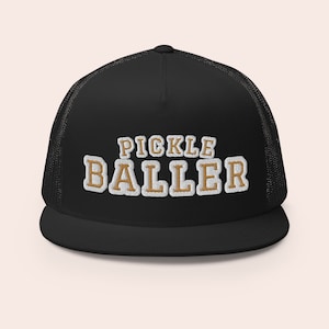 May include: Black trucker hat with a white mesh back. The hat has a gold embroidered design that says "Pickle Baller".