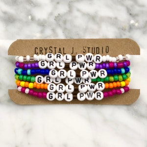 May include: A set of six colorful beaded bracelets with the words "GRL PWR" spelled out on each bracelet. The bracelets are arranged in a rainbow pattern, with each bracelet a different color.