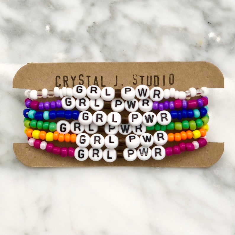 May include: A set of six colorful beaded bracelets with the words "GRL PWR" spelled out on each bracelet. The bracelets are arranged in a rainbow pattern, with each bracelet a different color.