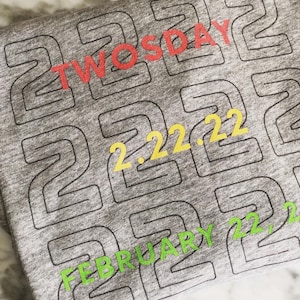 May include: Grey t-shirt with a repeating pattern of the number 2. The text "TWOSDAY" is printed in red and the text "FEBRUARY 22, 22" is printed in yellow and green.