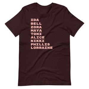 K&ouml;nnte beinhalten: Ein maroonfarbenes T-Shirt mit einer vertikalen Liste von Namen in rosa gedruckt: Ida Bell, Zora, Maya, Toni, Alice, Nikki, Phillis, Lorraine.