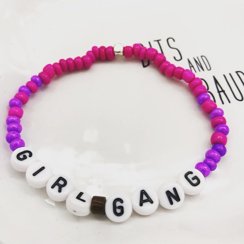 K&ouml;nnte beinhalten: Ein Armband mit Perlen, auf dem in wei&szlig;en Buchstaben "GIRL GANG" steht. Das Armband hat eine Kombination aus rosa, lila und silbernen Perlen und ist ein farbenfrohes Accessoire.