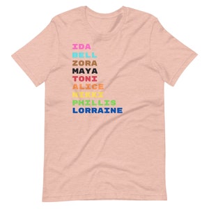 K&ouml;nnte beinhalten: Ein pinkfarbenes T-Shirt mit einem Regenbogen aus Namen auf der Vorderseite: Ida, Bell, Zora, Maya, Toni, Alice, Nikki, Phillis, Lorraine.
