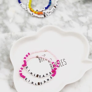 K&ouml;nnte beinhalten: Zwei Armb&auml;nder aus rosa und pinkfarbenen Perlen mit dem Schriftzug "GIRL GANG" aus wei&szlig;en Buchstabenperlen. Goldfarbene Akzente sind vorhanden. Weitere Armb&auml;nder mit bunten Perlen sind im Hintergrund zu sehen, auf einem wei&szlig;en, wolkenf&ouml;rmigen Teller.
