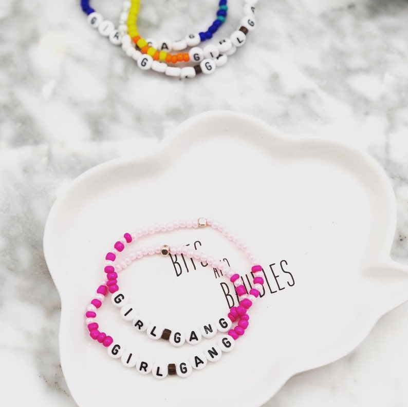 K&ouml;nnte beinhalten: Zwei Armb&auml;nder aus rosa und pinkfarbenen Perlen mit dem Schriftzug "GIRL GANG" aus wei&szlig;en Buchstabenperlen. Goldfarbene Akzente sind vorhanden. Weitere Armb&auml;nder mit bunten Perlen sind im Hintergrund zu sehen, auf einem wei&szlig;en, wolkenf&ouml;rmigen Teller.