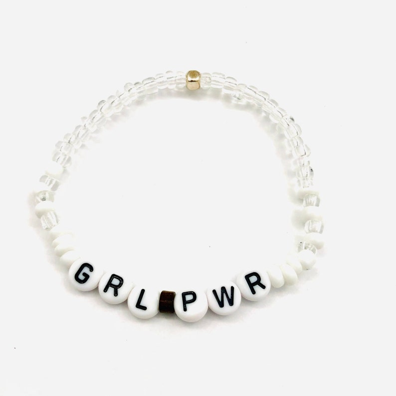 May include: White beaded bracelet with the word "GRL PWR" spelled out in black letters on the beads.