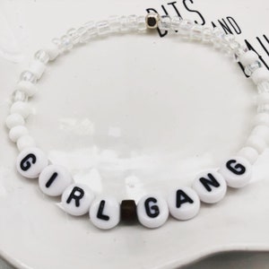 K&ouml;nnte beinhalten: Wei&szlig;es Perlenarmband mit den Worten "GIRL GANG" in schwarzen Buchstaben auf wei&szlig;en runden Perlen. Das Armband hat auch klare und wei&szlig;e Perlen. Eine kleine goldfarbene Perle ist sichtbar.