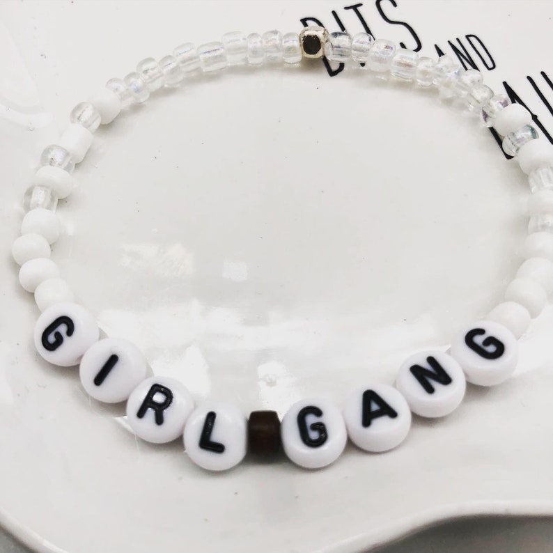 K&ouml;nnte beinhalten: Wei&szlig;es Perlenarmband mit den Worten "GIRL GANG" in schwarzen Buchstaben auf wei&szlig;en runden Perlen. Das Armband hat auch klare und wei&szlig;e Perlen. Eine kleine goldfarbene Perle ist sichtbar.