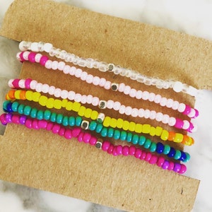 K&ouml;nnte beinhalten: Eine Sammlung farbenfroher Perlenarmb&auml;nder. Die Armb&auml;nder weisen eine Vielzahl von Farben auf, darunter Rosa, Gelb, T&uuml;rkis und Wei&szlig;. Jedes Armband hat kleine goldfarbene Perlen dazwischen. Die Armb&auml;nder werden auf einer braunen Kartonoberfl&auml;che pr&auml;sentiert.
