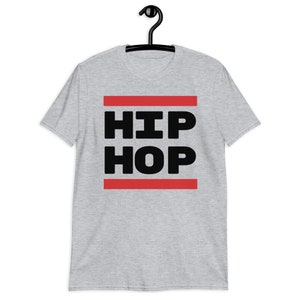 May include: A light grey t-shirt featuring the words "HIP HOP" in bold black capital letters, set between two red horizontal lines. The t-shirt is displayed on a black hanger.
