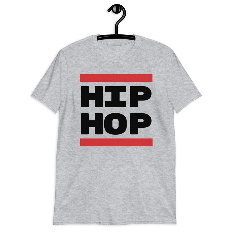 May include: A light grey t-shirt featuring the words "HIP HOP" in bold black capital letters, set between two red horizontal lines. The t-shirt is displayed on a black hanger.