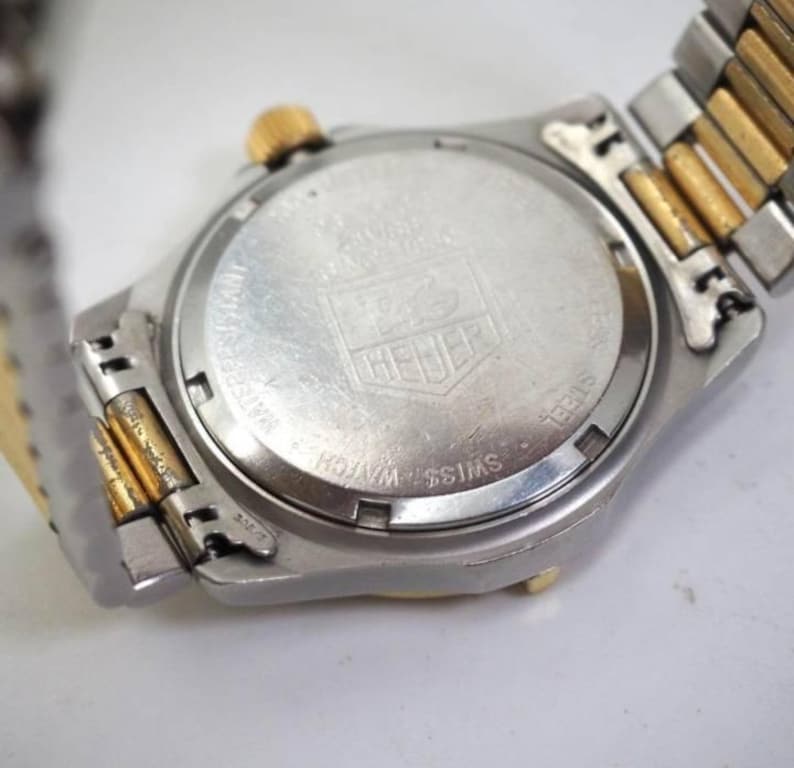 Pu&ograve; includere: Primo piano del retro di un orologio da polso argentato e dorato. Il quadrante dell'orologio &egrave; rotondo con finitura argentata e il marchio "HEUER" &egrave; visibile. Il cinturino &egrave; una combinazione di maglie argentate e dorate.