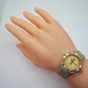 Pu&ograve; includere: Un orologio bicolore con lunetta dorata e cinturino argentato. Il quadrante dell'orologio &egrave; dorato con numeri e lancette nere. L'orologio &egrave; su un polso.