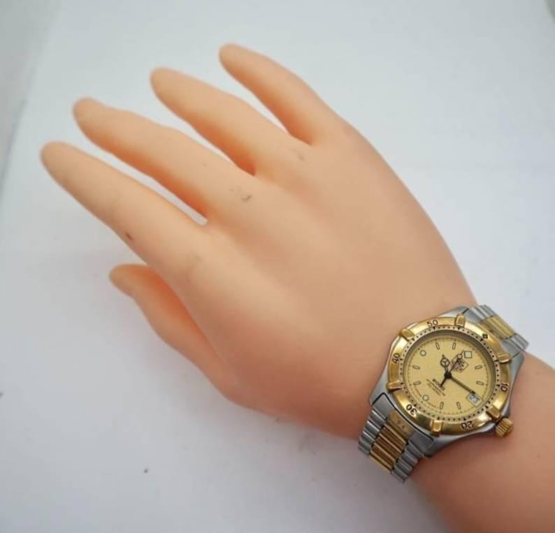 Pu&ograve; includere: Un orologio bicolore con lunetta dorata e cinturino argentato. Il quadrante dell'orologio &egrave; dorato con numeri e lancette nere. L'orologio &egrave; su un polso.