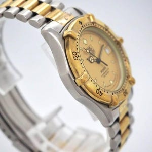 Pu&ograve; includere: Un orologio da polso bicolore con lunetta dorata e cinturino argentato. Il quadrante &egrave; color oro chiaro con lancette e indici neri. L'orologio &egrave; un TAG Heuer serie 2000.