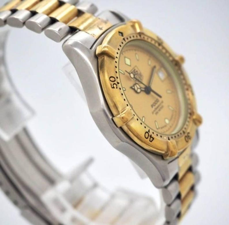 Pu&ograve; includere: Un orologio da polso bicolore con lunetta dorata e cinturino argentato. Il quadrante &egrave; color oro chiaro con lancette e indici neri. L'orologio &egrave; un TAG Heuer serie 2000.