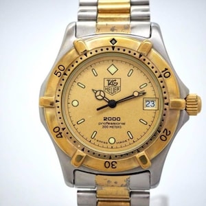 Pu&ograve; includere: Un orologio TAG Heuer 2000 Professional in oro e argento. L'orologio presenta un quadrante dorato con lancette e indici neri, un datario a ore 3 e una lunetta dorata. L'orologio &egrave; impermeabile fino a 200 metri e ha un bracciale in argento e oro.