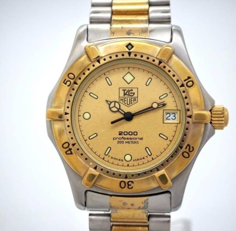 Pu&ograve; includere: Un orologio TAG Heuer 2000 Professional in oro e argento. L'orologio presenta un quadrante dorato con lancette e indici neri, un datario a ore 3 e una lunetta dorata. L'orologio &egrave; impermeabile fino a 200 metri e ha un bracciale in argento e oro.