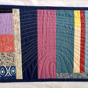 May include: A long, narrow quilt with a patchwork design. The quilt is made up of various shades of blue, red, yellow, and purple fabric. The quilt is quilted with a variety of patterns, including swirls, lines, and dots.